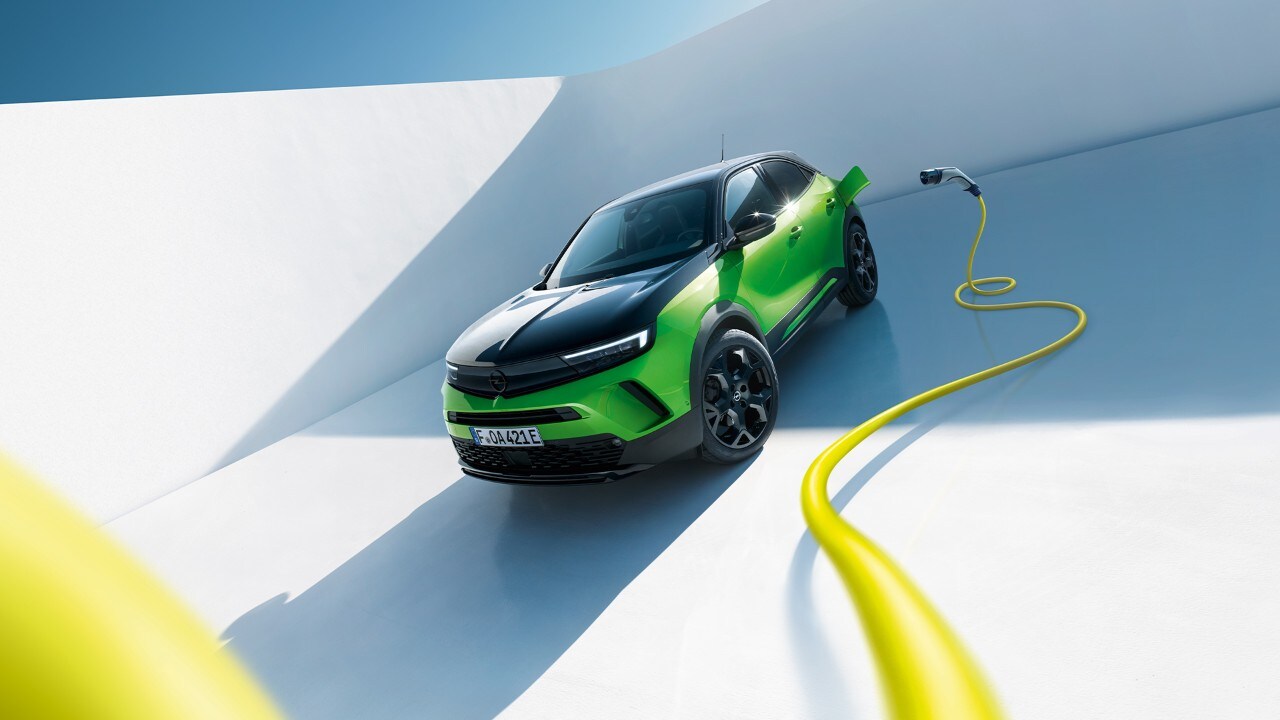 Vista lateral delantera de un Opel Mokka Eléctrico verde con llantas de aleación negras y techo con un cable de carga EV amarillo.